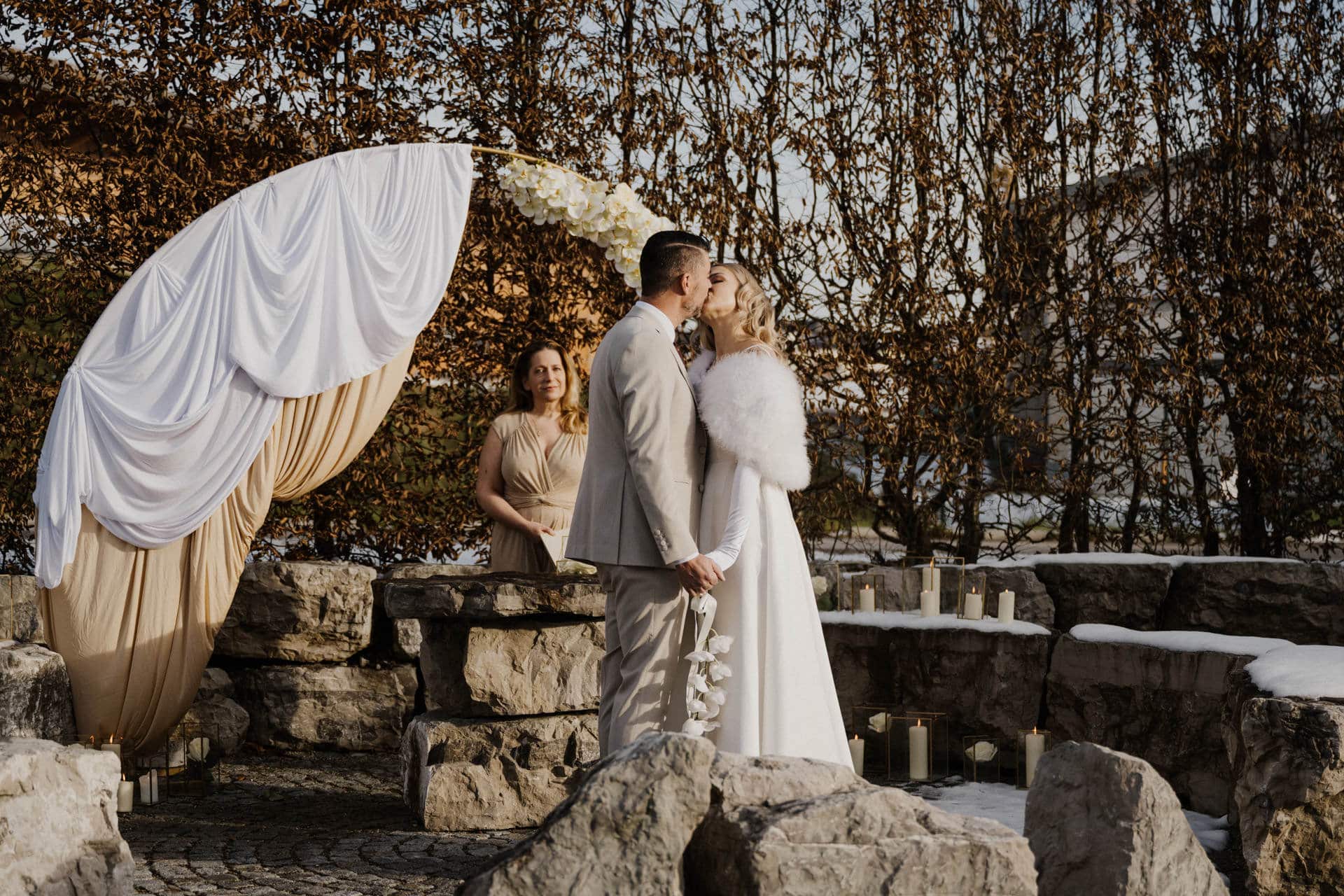 Braut und Bräutigam küssen sich während einer Winterhochzeit im Freien vor drapierten Stoffen und Steintreppen, mit einem Trauzeugen und einer Reihe von Kerzen im Hintergrund. Schnee ist auf den Felsen zu sehen.