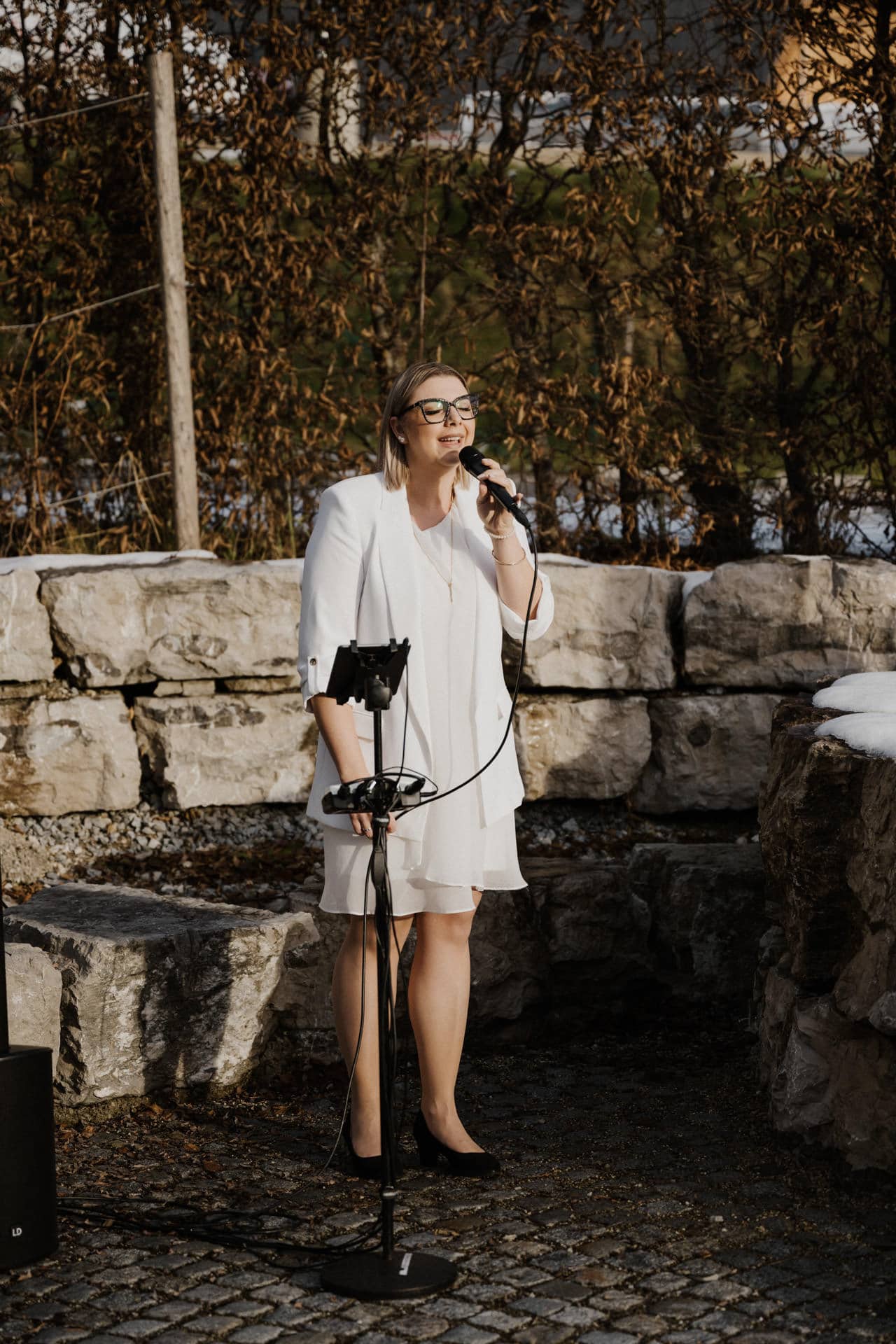 Eine Frau in weißem Kleid und Blazer singt im Freien in ein Mikrofon. Sie steht auf einem Steinpflaster mit großen Steinblöcken und spärlichem braunem Laub im Hintergrund. Sie hält das Mikrofon mit einer Hand.