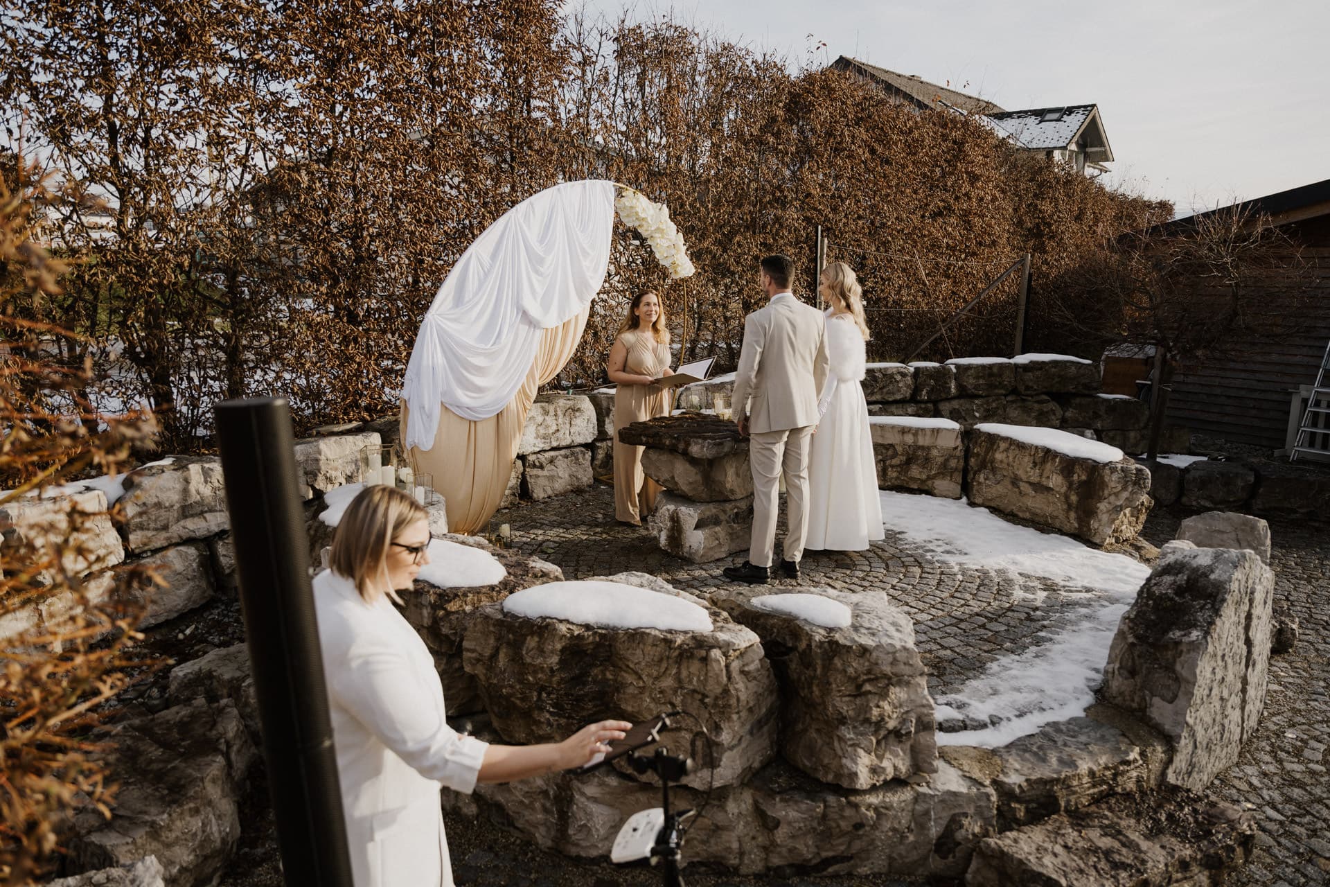 Eine kleine Hochzeitszeremonie im Freien mit einem Paar, das vor einem Zeremonienmeister in einem Steinkreis steht, umgeben von schneebedeckten Felsen, während eine andere Person im Vordergrund eine Kamera einstellt.