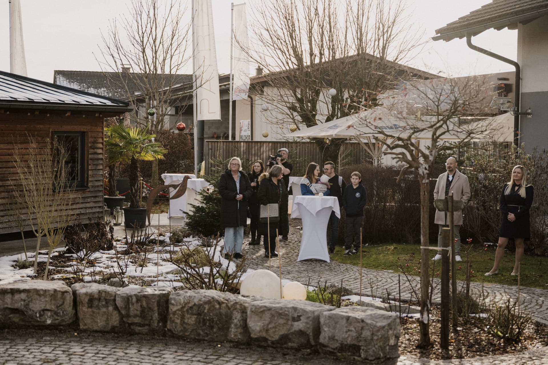 Eine Gruppe von Personen, einige mit Getränken in der Hand, steht im Freien in einem Garten mit kahlen Bäumen und Schneeflecken. Sie sind warm gekleidet und scheinen an einem kleinen geselligen Beisammensein in der Nähe mehrerer Häuser teilzunehmen.