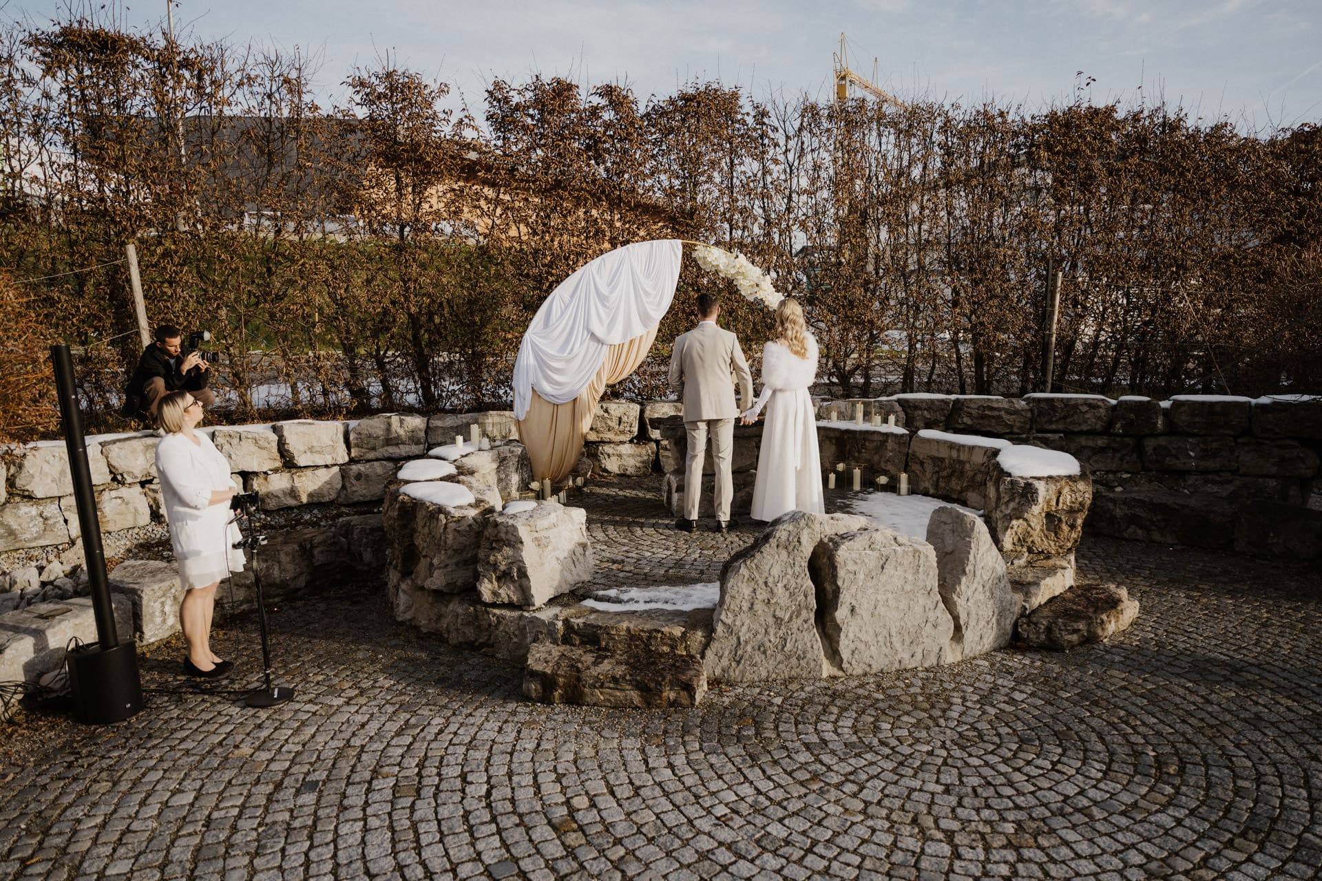 Ein Paar steht gemeinsam vor einem mit weißem Stoff und Blumen geschmückten Steinaltar im Freien. Ein Fotograf hält den Moment fest, während eine weitere Person in der Nähe steht. Die Szene ist von blattlosen Bäumen und Kopfsteinpflaster umgeben.