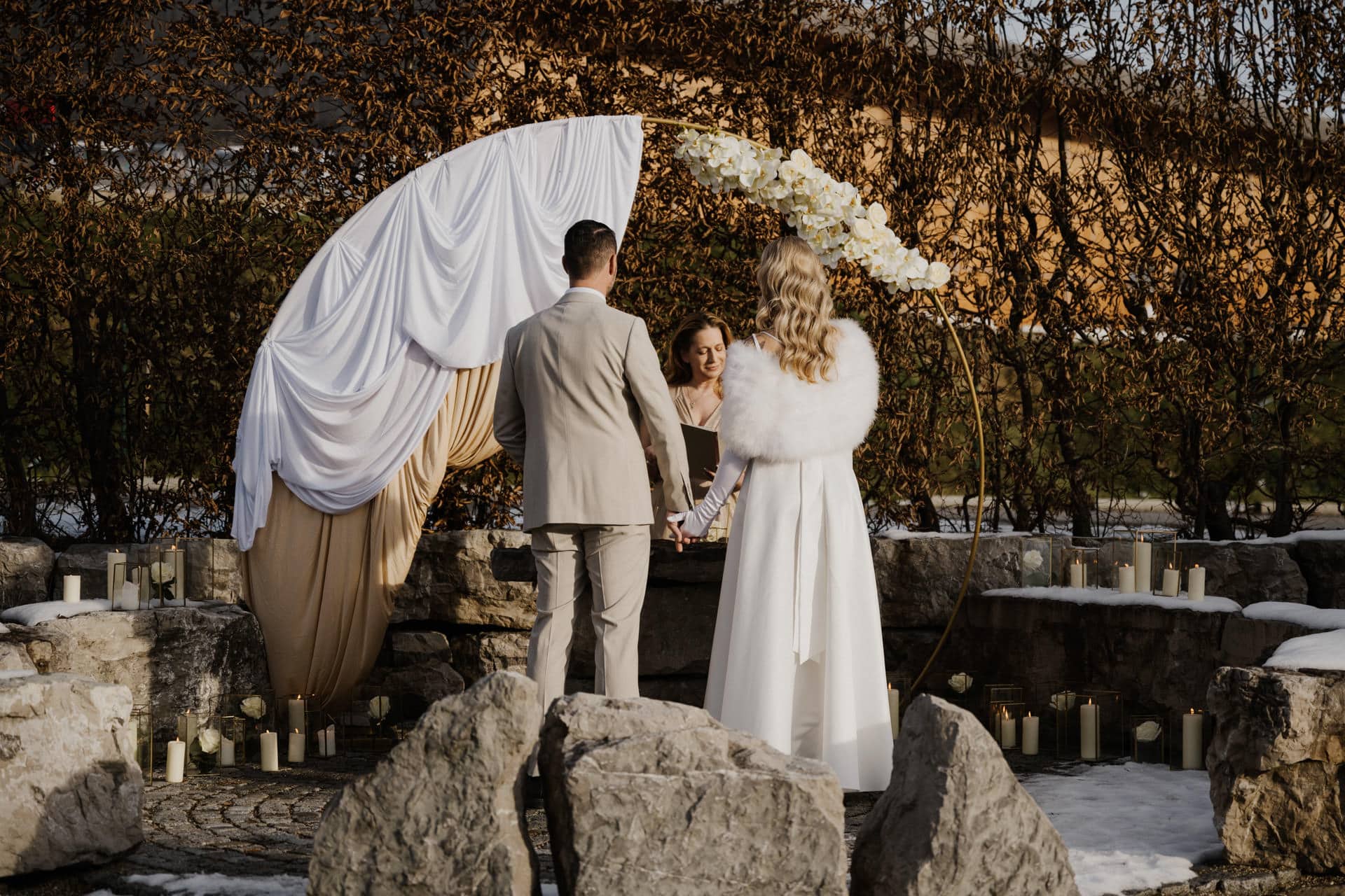 Ein formell gekleidetes Paar hält sich während einer Hochzeitszeremonie im Freien an den Händen, während es einem Geistlichen gegenübersteht. Sie stehen vor einem mit Blumen drapierten Bogen, umgeben von Steinen und Grünzeug, mit Kerzen und einigen Schneeflecken im Blick.