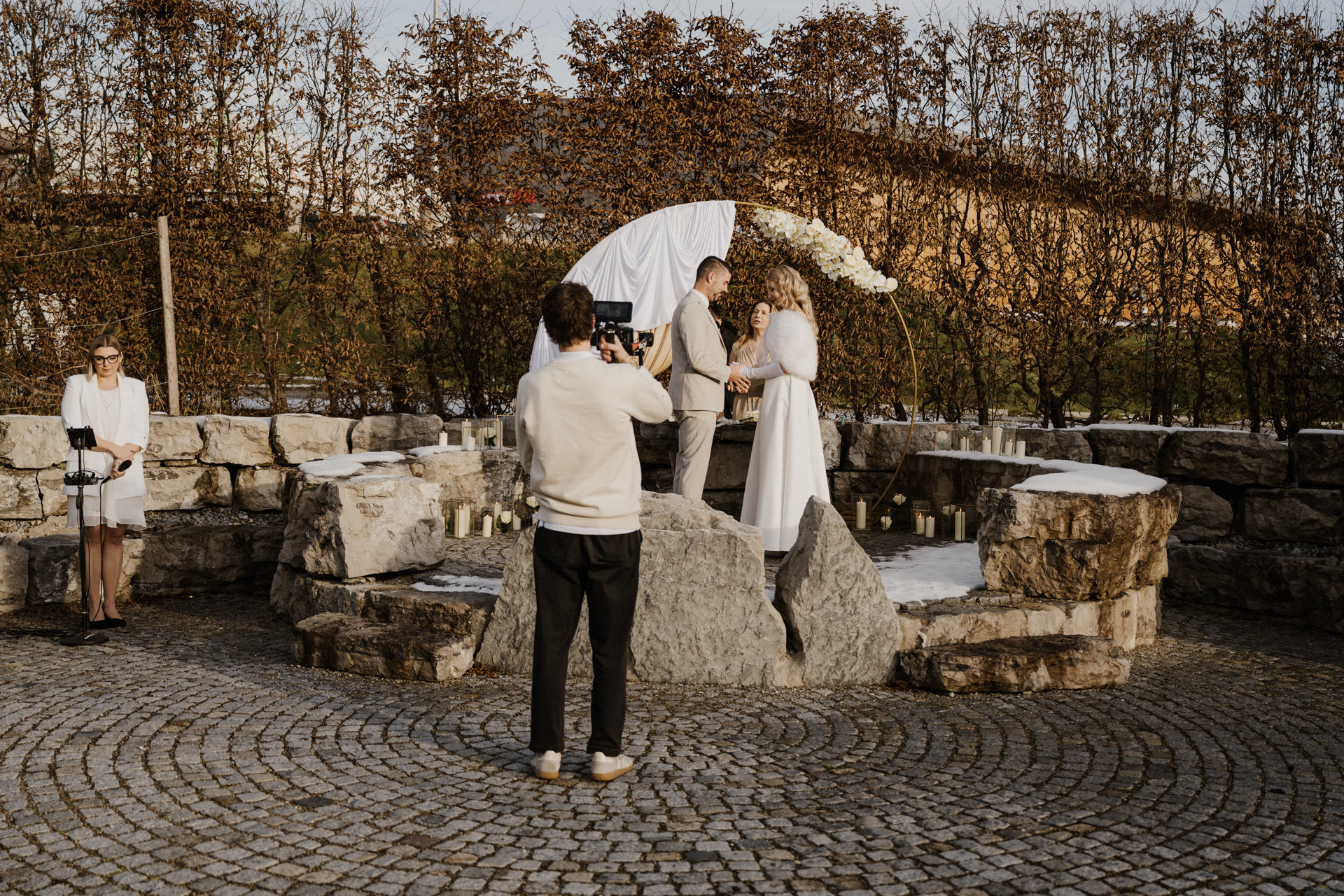 Ein Paar steht auf einer großen Steintreppe und gibt sich unter einem weißen Blumenbogen im Freien das Eheversprechen, während eine Person sie fotografiert und eine weitere Person an der Seite steht und die Papiere hält. Die Kulisse ist von blattlosen Bäumen und Steinmauern umgeben.