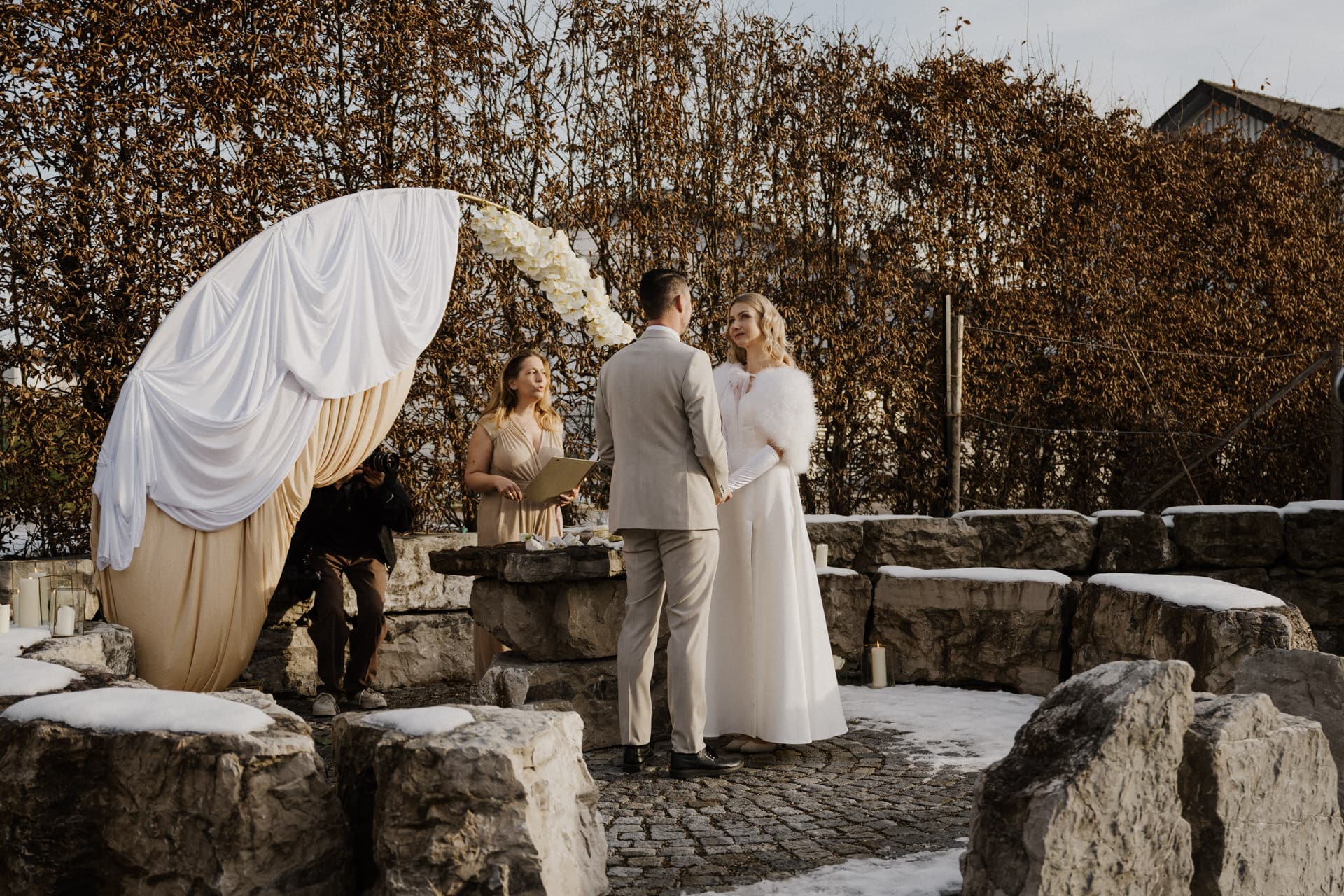Ein Paar steht bei einer Trauung im Freien Hand in Hand, dem Trauredner gegenüber. Sie sind von Steinstrukturen und Schnee umgeben, während ein Fotograf den Moment in der Nähe eines drapierten Bogens festhält.