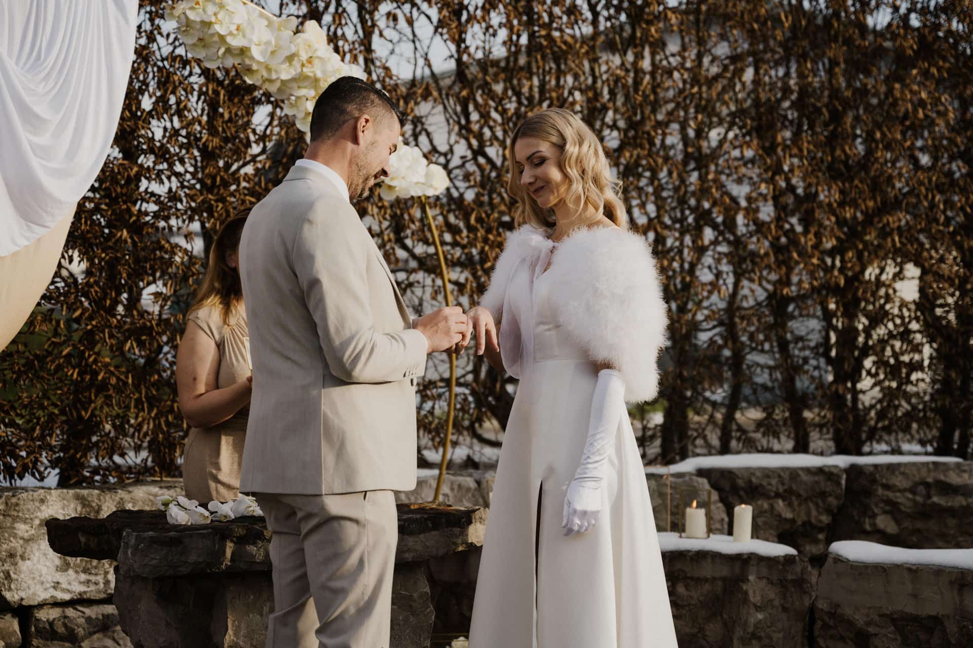 Ein Paar steht während einer Hochzeitszeremonie im Freien. Der Bräutigam steckt einen Ring an den Finger der Braut. Die Braut trägt ein weißes Kleid mit Handschuhen und einem Pelzschal, während der Bräutigam einen hellen Anzug trägt.