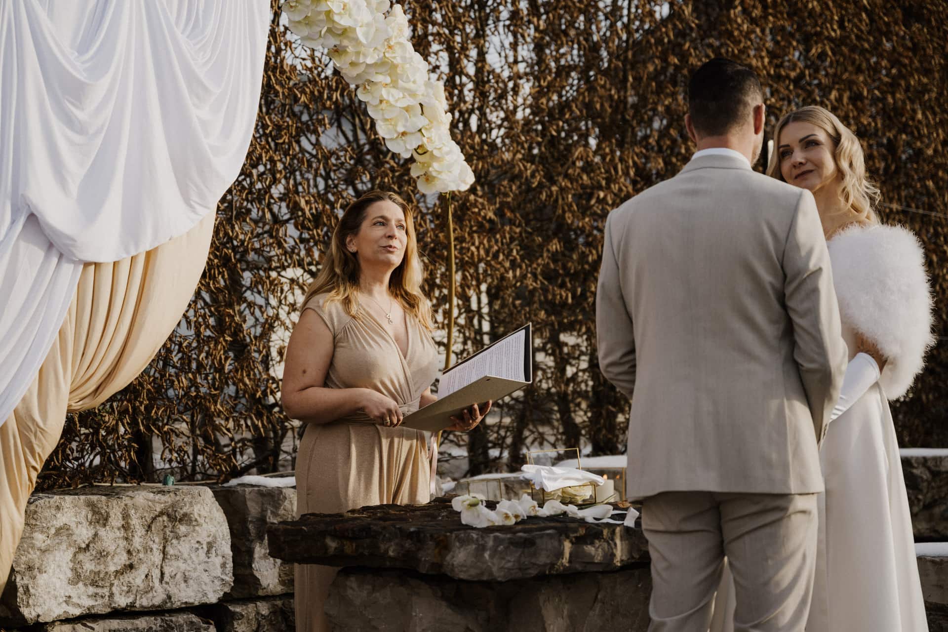 Eine Hochzeitszeremonie im Freien: Ein Zeremonienmeister steht an einem Steintisch mit Dokumenten, während sich Braut und Bräutigam gegenüberstehen und die Hände halten. Die Braut trägt ein weißes Kleid und ein Kopftuch, der Bräutigam einen hellen Anzug. Drapierte Stoffe und Blumen schmücken die Szene.