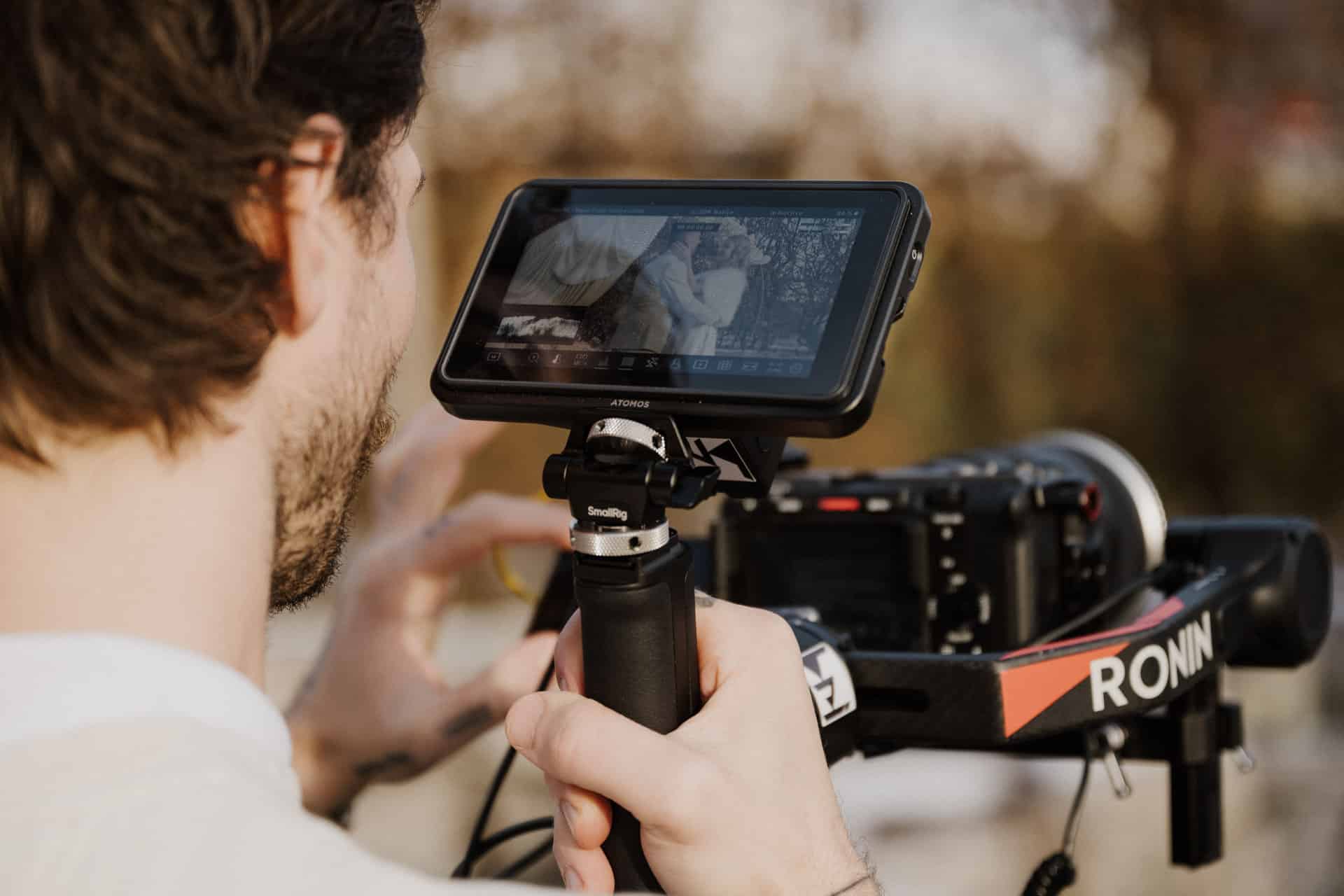 Eine Person bedient eine Videokamera, die auf einem Ronin-Stabilisator montiert ist, und sieht sich die Aufnahmen eines Paares auf dem angeschlossenen Bildschirm im Freien an.