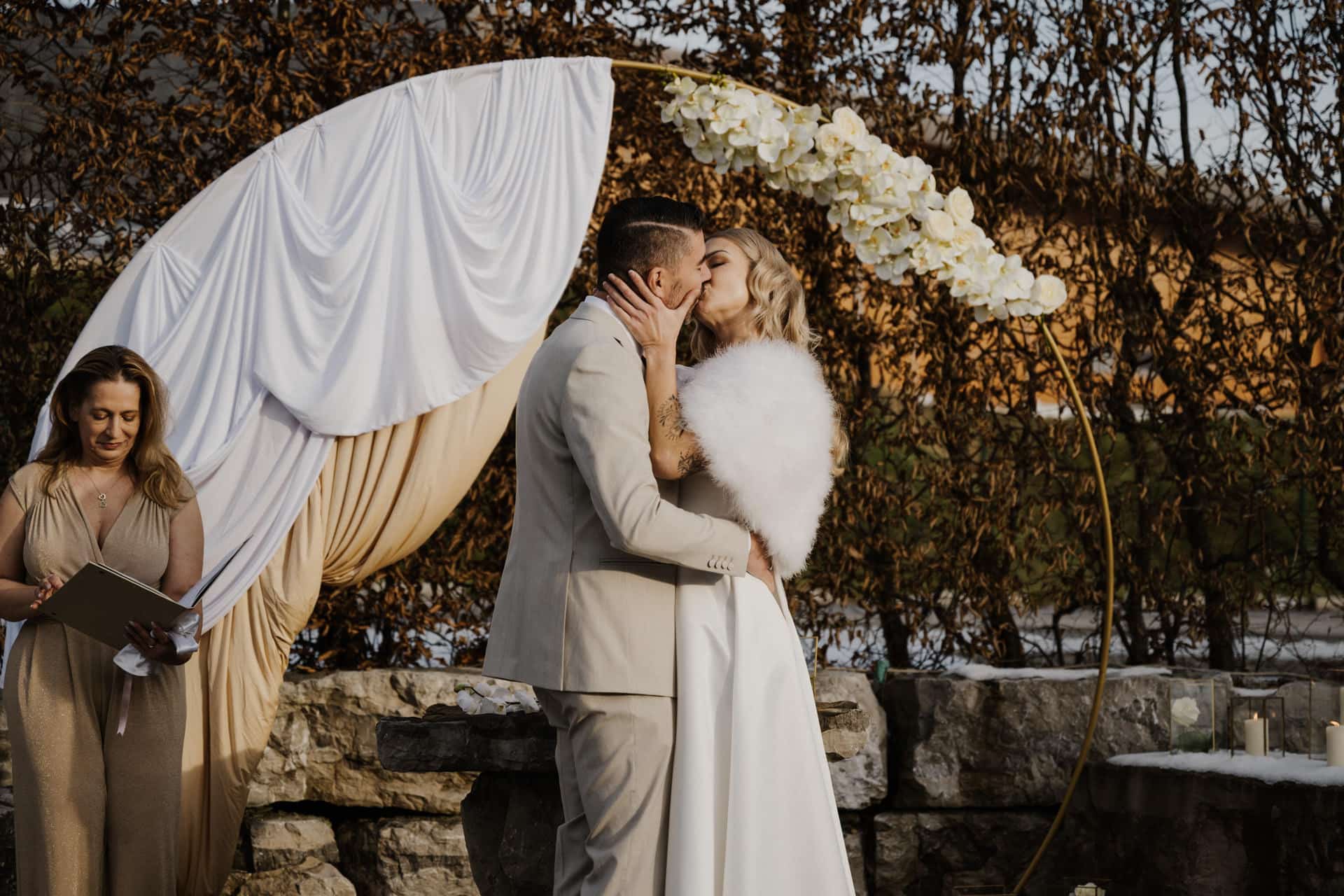 Eine Braut und ein Bräutigam küssen sich unter einem runden, mit weißem Stoff und Blumen geschmückten Hochzeitsbogen, während eine Frau in einem goldenen Kleid daneben steht und ein Buch hält. Sie befinden sich im Freien, mit Grünpflanzen und Steinmauern im Hintergrund.