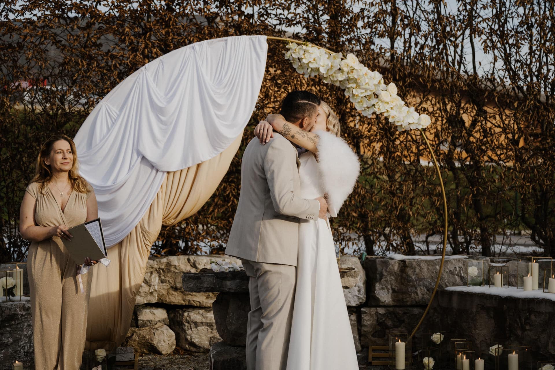Braut und Bräutigam küssen sich bei einer Hochzeitszeremonie im Freien, umrahmt von einem Bogen mit weißem Stoff und Blumen. In der Nähe steht eine Frau mit einem Buch in der Hand, und auf den Steinstufen sind Kerzen aufgestellt.
