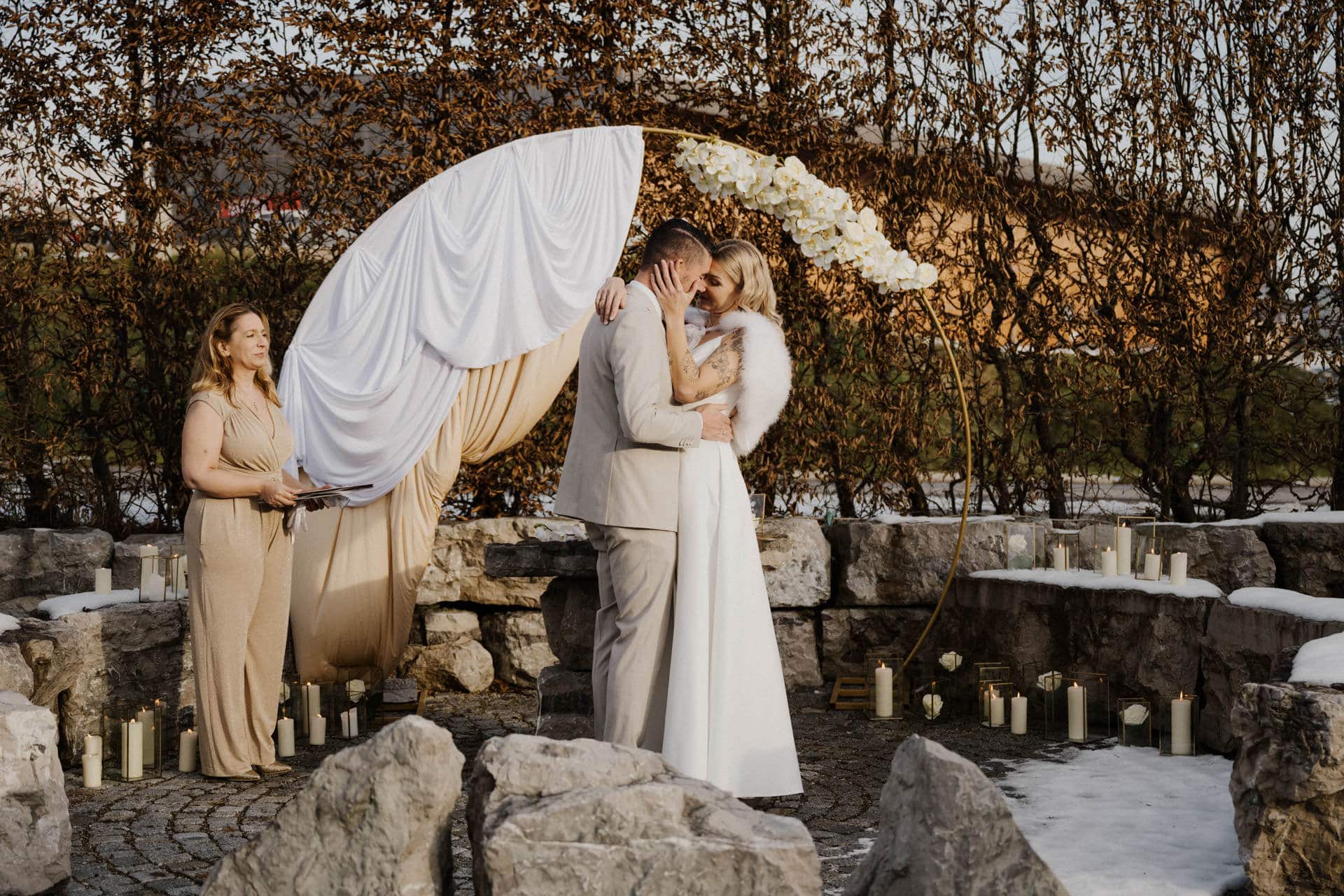 Ein frisch verheiratetes Paar umarmt und küsst sich im Freien vor einem runden, mit weißem Stoff und Blumen drapierten Hochzeitsbogen, während eine Frau in einem beigen Kleid daneben steht. Die Szene ist von Kerzen und Steinen umgeben; auf dem Boden ist Schnee zu sehen.