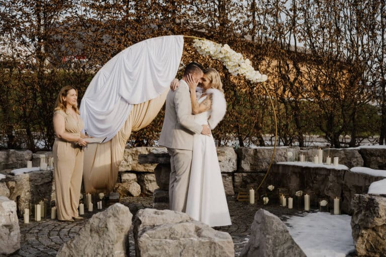 Bei einer Winterhochzeit im Freien küssen sich Braut und Bräutigam unter einem mit Blumen geschmückten weißen Stoffbogen. Eine Brautjungfer in einem goldenen Kleid steht daneben, während Kerzen in Glashaltern den Steinaltar umgeben.