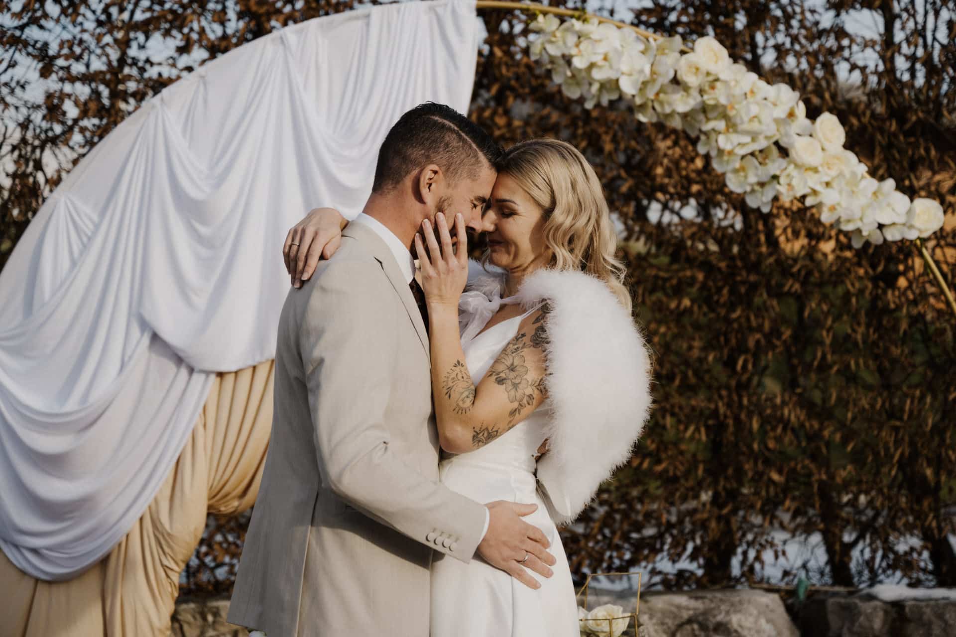 Eine Braut und ein Bräutigam umarmen sich und lächeln bei einer Hochzeit im Freien eng beieinander, während sie vor einem weiß-gold drapierten Bogen mit weißen Blumen stehen. Die Braut trägt eine Pelzstola und hat eine sichtbare Armtätowierung.