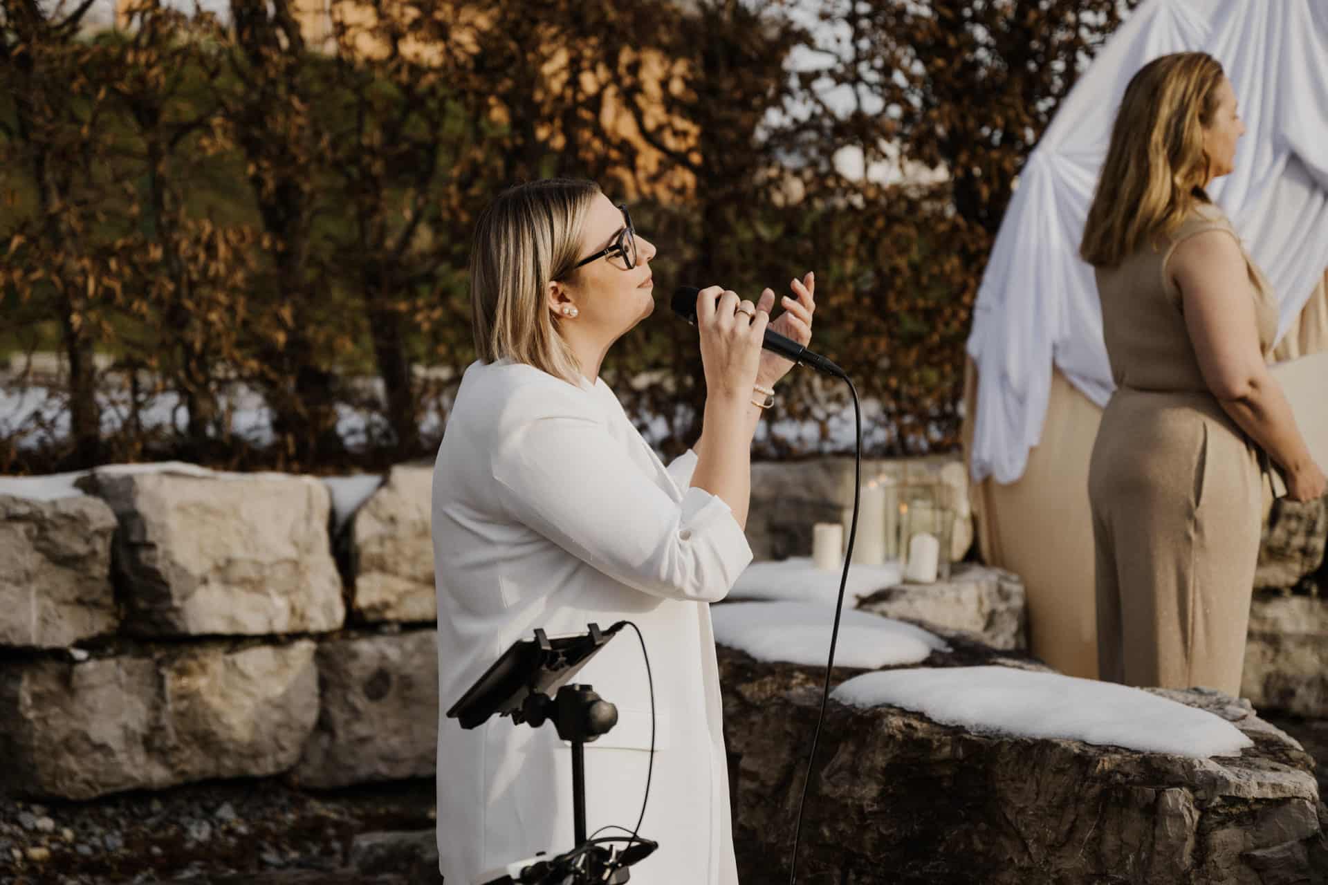 Eine Frau in einem weißen Blazer hält ein Mikrofon in der Hand und singt im Freien in der Nähe großer Steinblöcke, während eine andere Frau in einem beigen Kleid in der Nähe steht. Auf den Felsen liegt Schnee und im Hintergrund ist weißer Stoff drapiert.