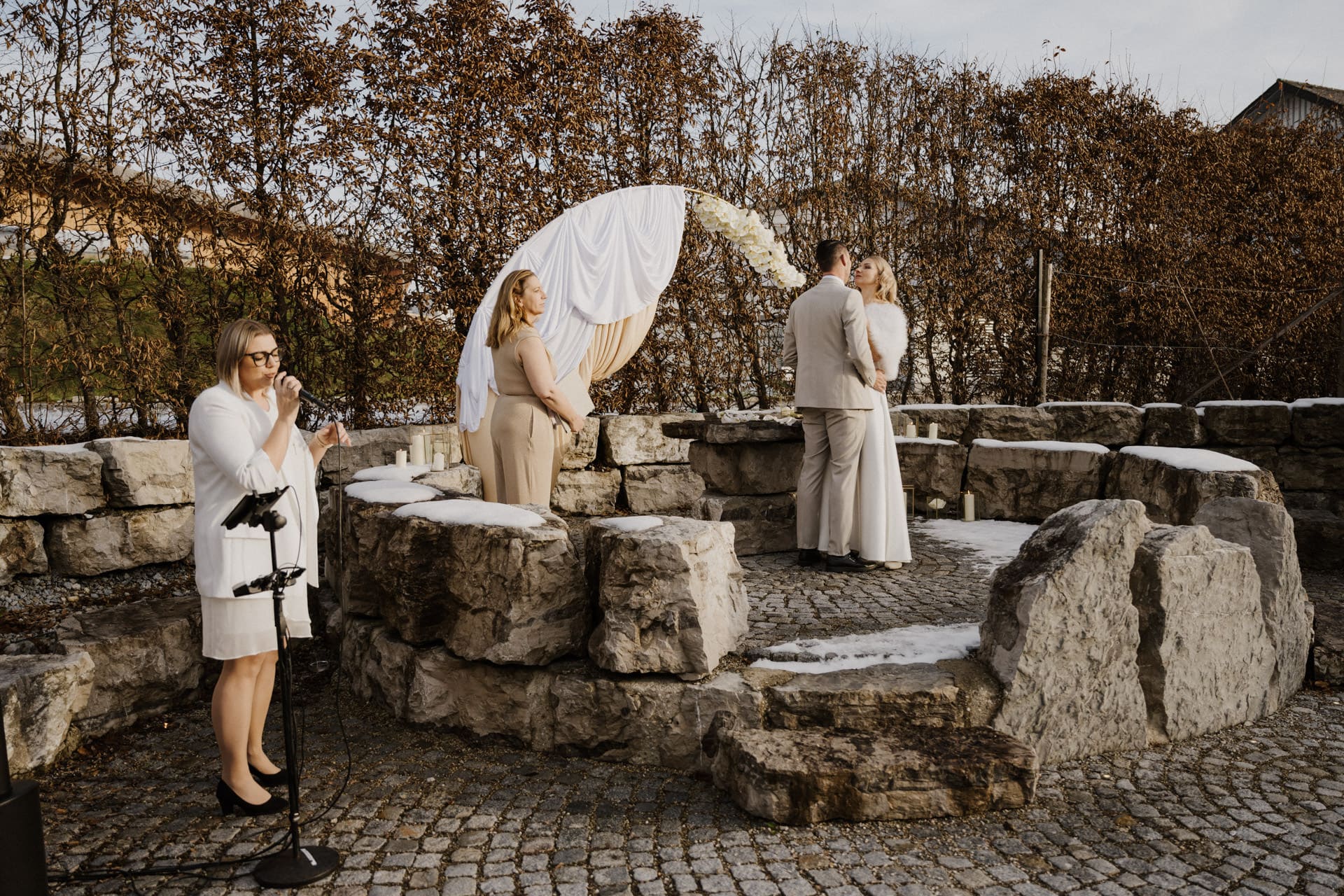 Eine Trauung im Freien auf einer Steintreppe, wobei sich das Paar unter einem weißen Blumenbogen gegenübersteht. Zwei Frauen stehen in der Nähe; eine singt oder spricht in ein Mikrofon, die andere beobachtet das Paar.