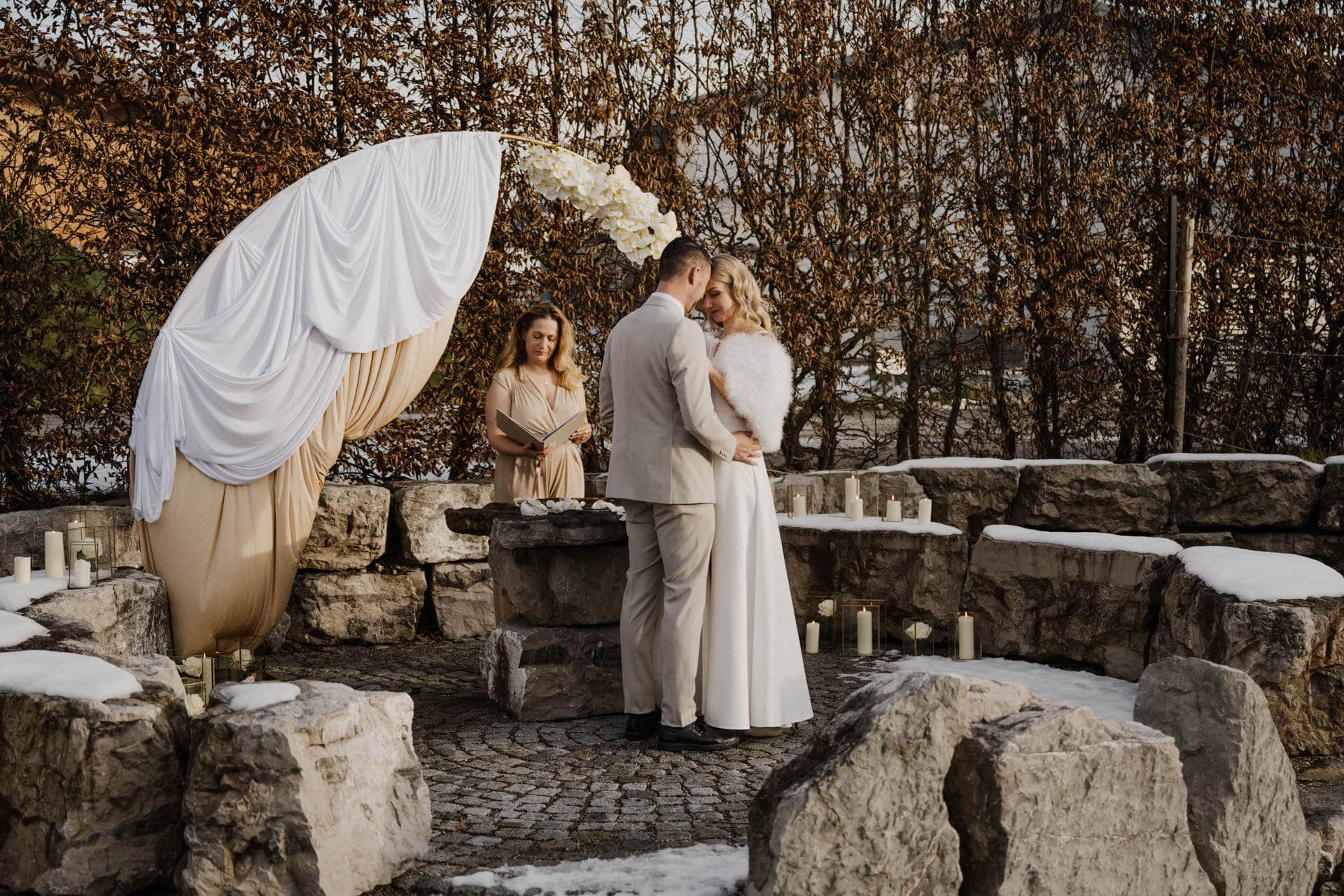 Ein Paar in Hochzeitskleidung steht eng beieinander in einem Steinkreis im Freien, umgeben von Schnee und Kerzen, während eine Trauzeugin in einem beigen Kleid hinter ihnen in der Nähe eines weiß verhüllten Bogens steht.
