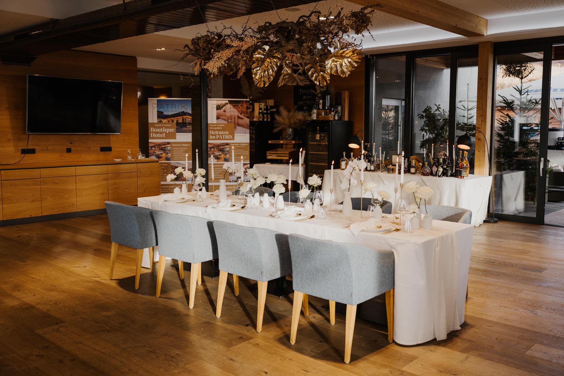 Ein modernes Esszimmer mit einem langen Tisch, der für ein formelles Essen gedeckt ist, dekoriert mit weißer Bettwäsche, Kerzen und eleganten Blumenarrangements. Um den Tisch herum stehen sechs blaue Stühle, über denen große goldene Blätter hängen.