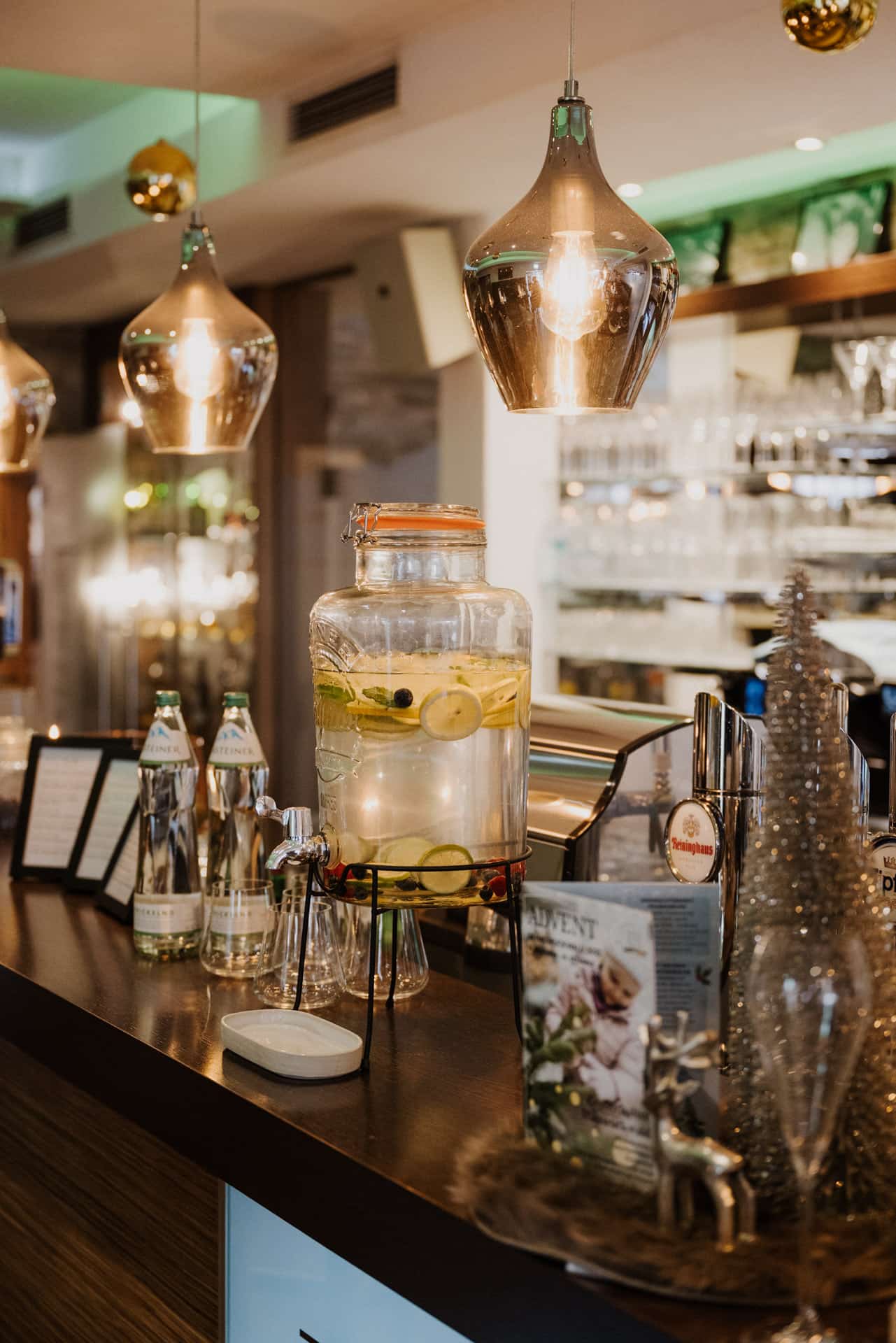 Ein großer Glasspender, gefüllt mit Wasser, Zitrone und Gurkenscheiben, steht auf einem Bartresen, umgeben von Flaschen, Gläsern und festlichen Dekorationen mit warmen Hängelampen über dem Kopf.