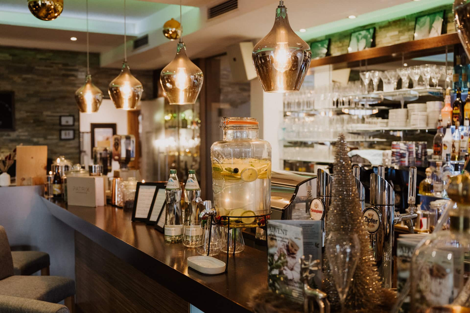 Eine moderne Bar mit goldenen Hängelampen, einem mit Flaschen bestückten Tresen, einem großen Glasspender mit Zitronen- und Gurkenwasser, Barhockern und Regalen mit Glaswaren und Flaschen im Hintergrund.