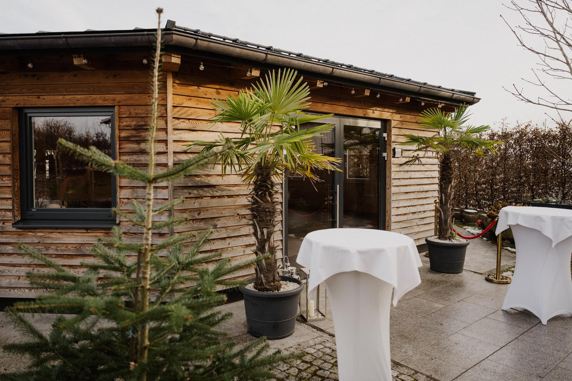 Ein Holzgebäude mit schwarz gerahmten Fenstern und einer Glastür ist umgeben von Palmen und weiß gedeckten Tischen auf einer gepflasterten Terrasse.