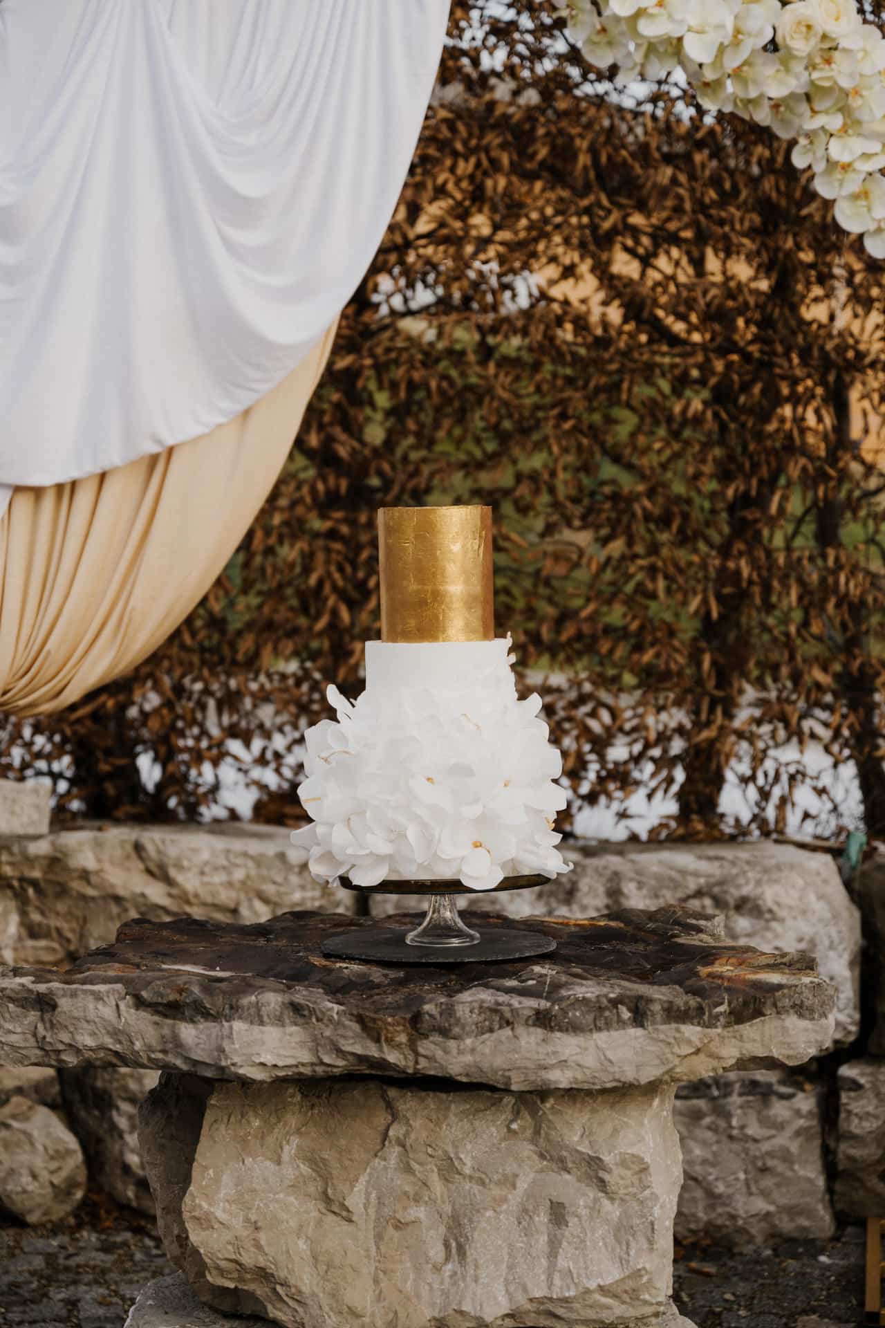 Eine zweistöckige Torte mit einer goldenen Oberschicht und einer mit weißen Blumen verzierten Unterschicht, die auf einem Glasständer auf einem Steintisch im Freien steht, mit drapiertem Stoff und Laub im Hintergrund.