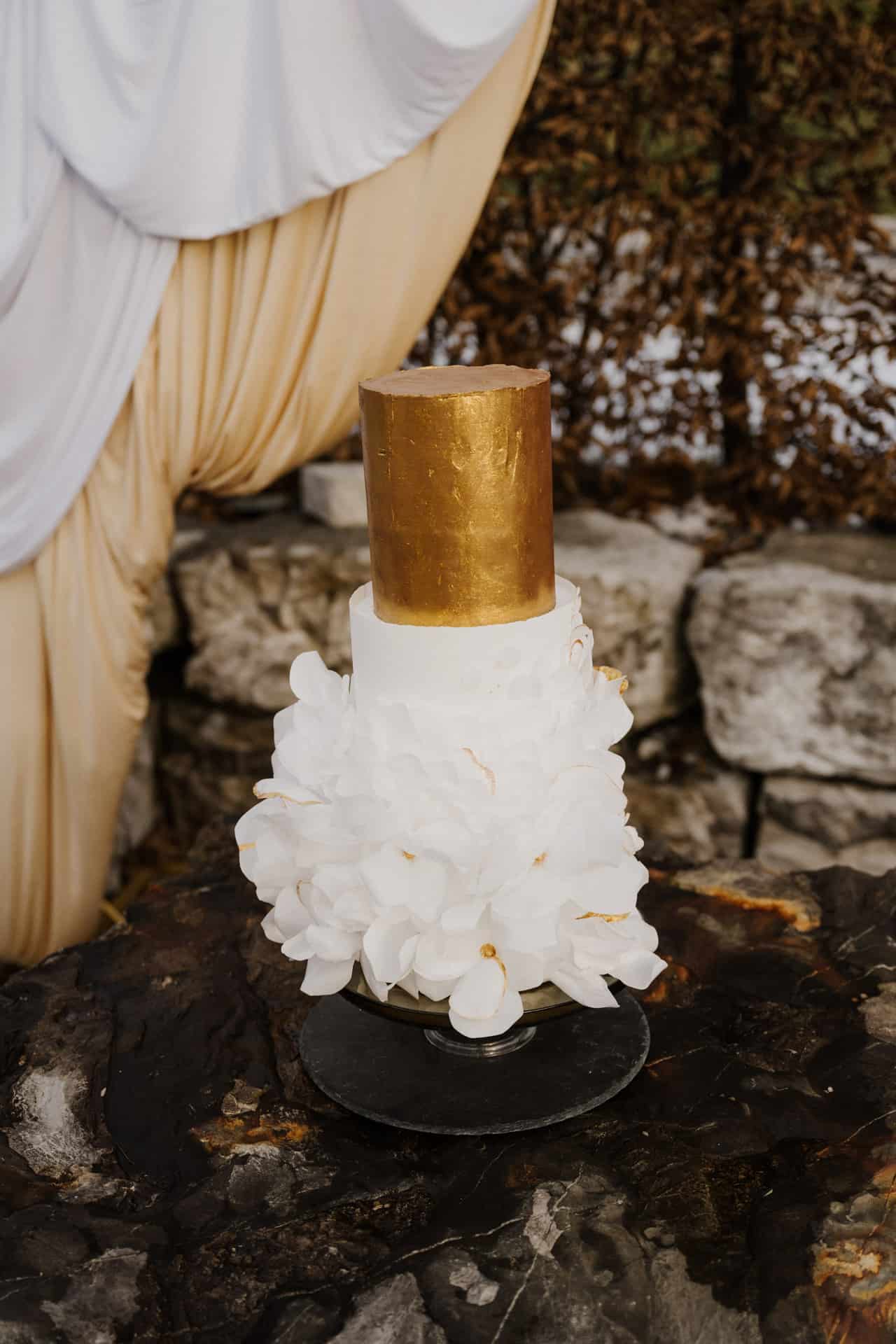 Eine zweistöckige Torte mit einer goldenen zylindrischen Oberschicht und einer weißen Unterschicht, die mit großen weißen Blumendekorationen verziert ist, steht auf einer dunklen Steinfläche mit drapiertem Stoff und Felsen im Hintergrund.