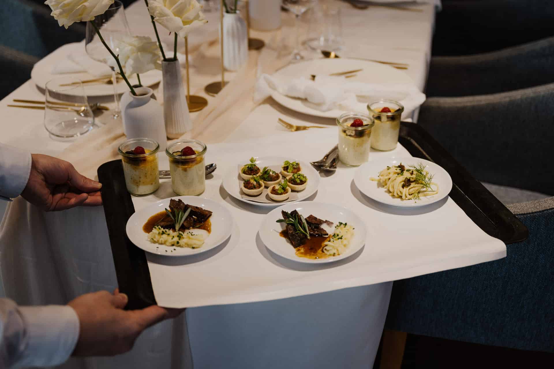 Eine Person serviert an einem elegant gedeckten Tisch mit weißen Blumen und goldenen Utensilien ein Tablett mit Gourmetgerichten, darunter Fleisch mit Kartoffelpüree, Pasta, Vorspeisen und Desserttassen.