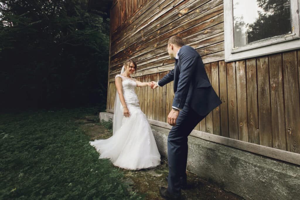 Eine Braut in einem weißen Kleid und ein Bräutigam in einem dunklen Anzug, inspiriert von den neuesten Hochzeitstrends, halten sich an den Händen und lächeln sich an, während sie draußen neben einem rustikalen Holzgebäude stehen, umgeben von Grün.