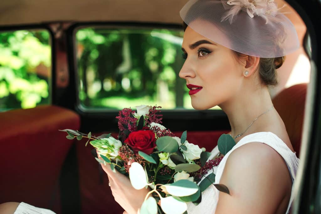 Eine Braut in weißem Kleid und Schleier, die die neuesten Hochzeitstrends aufgreift, hält einen Strauß roter und weißer Blumen, während sie auf dem Rücksitz eines Oldtimers sitzt und nachdenklich aus dem Fenster schaut.