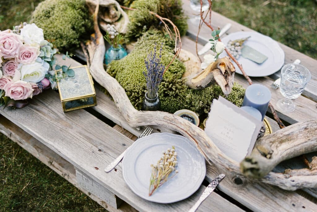 Eine rustikale Tischdekoration im Freien, inspiriert von Hochzeitstrends, mit Treibholz, Moos, Lavendel, rosa Rosen, Kerzen, Gläsern, Tellern und Besteck auf einer Holzpalette sorgt für eine natürliche und doch elegante Atmosphäre.