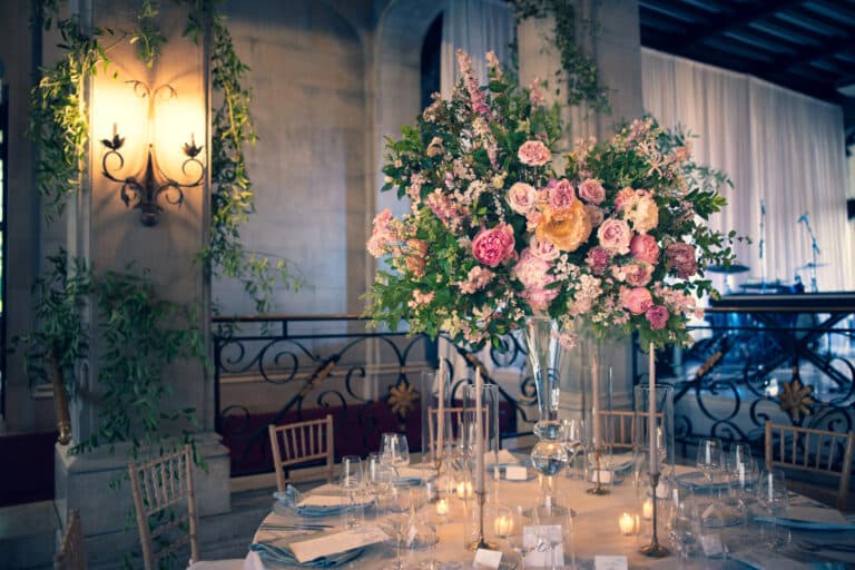 Eleganter runder Tisch für eine Veranstaltung auf einer Hochzeitsmesse, mit Gläsern, Tellern, Kerzen und einem großen floralen Tafelaufsatz mit rosa und gelben Blumen in einem großen, gedämpft beleuchteten Raum mit kunstvollem Dekor.