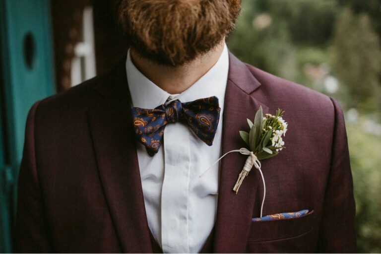 Ein Mann in einer weinroten Anzugsjacke mit einer Paisley-Fliege und einem passenden Einstecktuch, der ein weißes Hemd trägt. Eine kleine Blumenboutonniere ist an sein Revers geheftet. Der Hintergrund besteht aus unscharfem Grün.