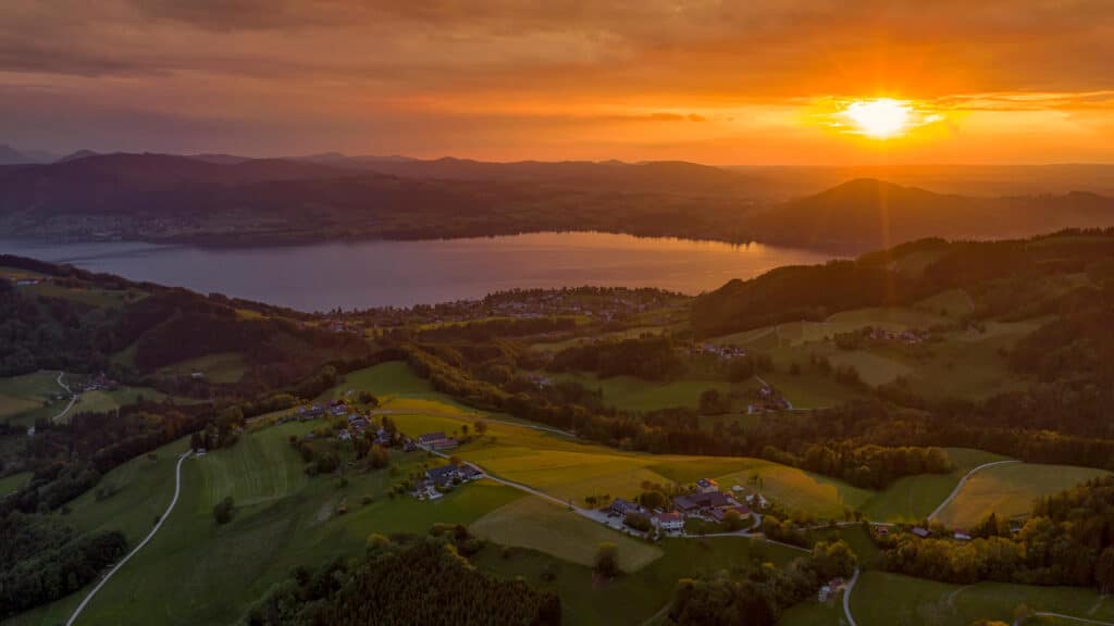 Ein strahlender Sonnenuntergang taucht einen ruhigen See in orangefarbenes Licht, der von sanften grünen Hügeln, vereinzelten Häusern und gewundenen Straßen umgeben ist, während am Horizont in der Ferne Berge zu sehen sind.