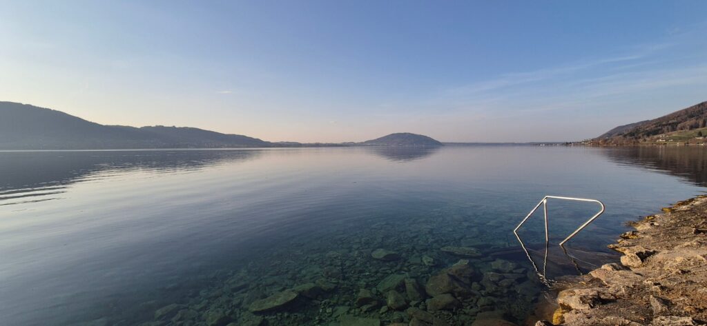 Ein ruhiger, klarer See mit felsigem Ufer, einer Metallleiter, die ins Wasser führt, fernen Hügeln und dem blauen Himmel, der sich auf der Wasseroberfläche spiegelt.