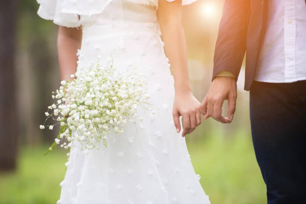 Eine Braut in einem weißen Kleid hält einen Strauß Schleierkraut in den Händen und spaziert Hand in Hand mit ihrem Partner im Freien, während sie das sanfte Sonnenlicht ihrer Sommerhochzeit genießt.