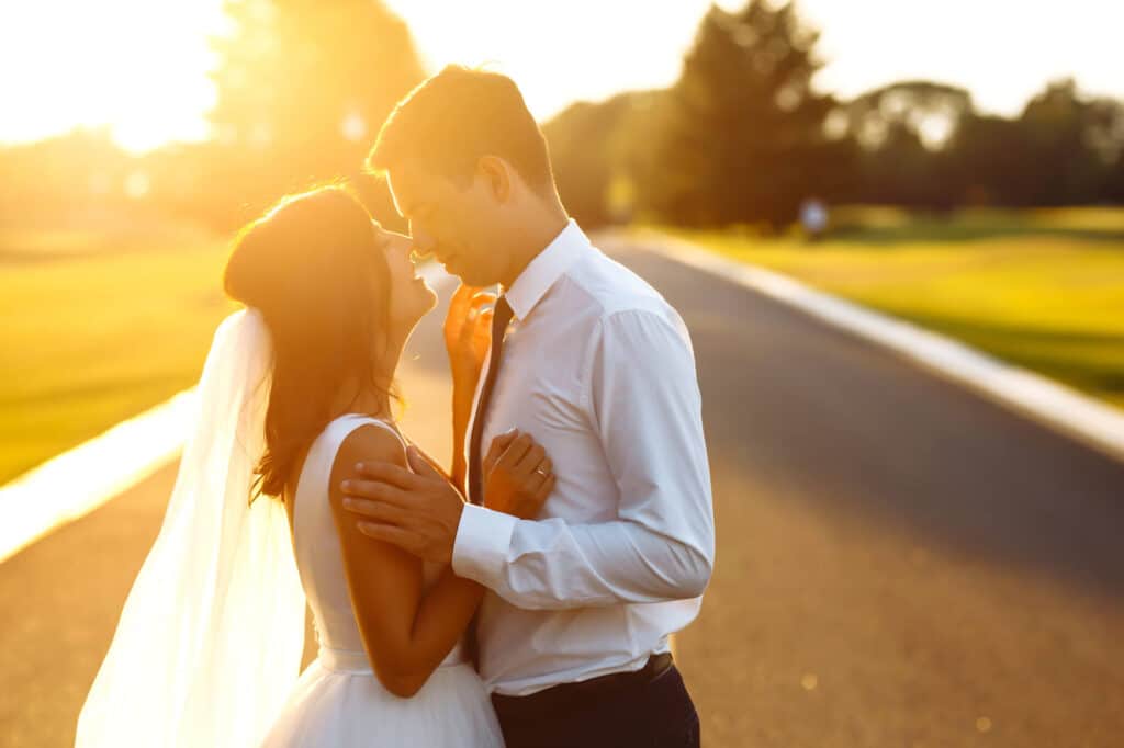 Ein Brautpaar umarmt sich während seiner Sommerhochzeit auf einer sonnenbeschienenen Straße, blickt sich tief in die Augen, während im Hintergrund Bäume und ein strahlender Sonnenuntergang zu sehen sind.