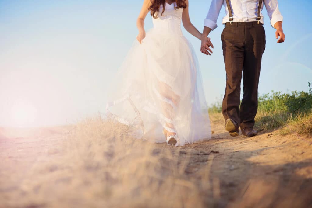 Eine Braut in einem weißen Kleid und ein Bräutigam mit Hosenträgern gehen Hand in Hand einen sonnendurchfluteten Weg entlang, der zu beiden Seiten von trockenem Gras gesäumt ist – ein Bild, das die unbeschwerte Romantik einer Sommerhochzeit einfängt.