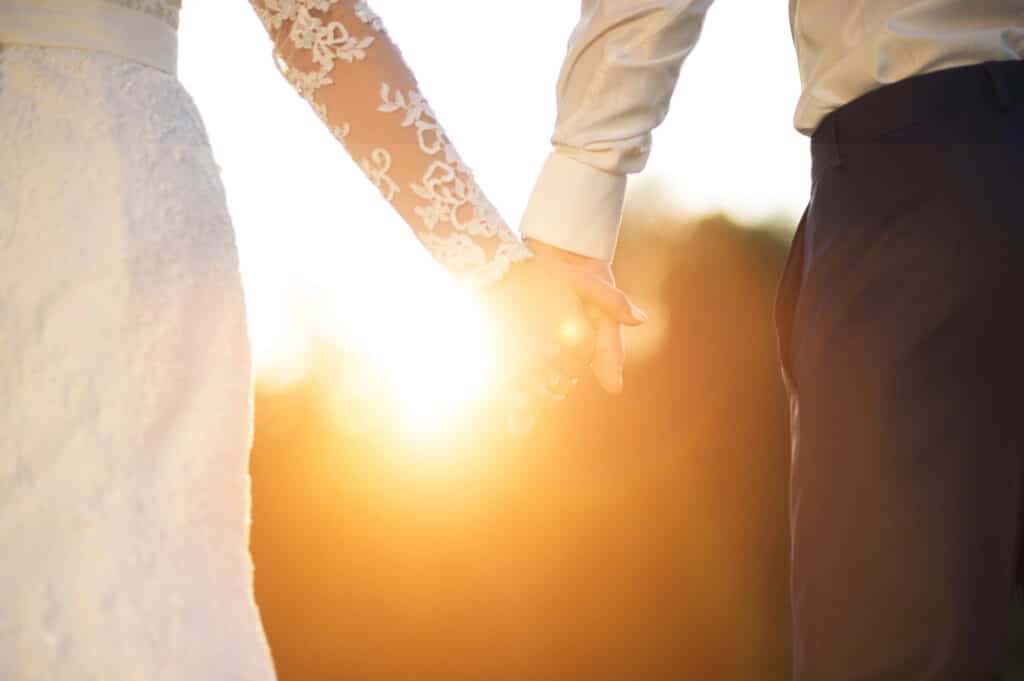 Ein Brautpaar hält sich bei Sonnenuntergang im Freien während seiner Sommerhochzeit an den Händen, während das Sonnenlicht durch ihre Finger scheint. Die Braut trägt ein Kleid mit Spitzenärmeln, und der Bräutigam trägt ein weißes Hemd und eine dunkle Hose.
