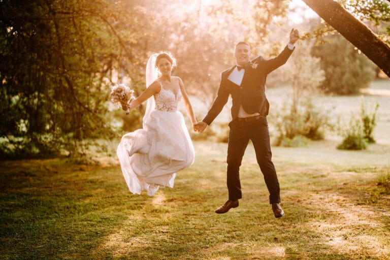 Ein Brautpaar, das sich an den Händen hält, springt während seiner Sommerhochzeit im Freien fröhlich in die Luft. Die Braut trägt ein weißes Kleid und hält einen Blumenstrauß in der Hand, während der Bräutigam einen schwarzen Anzug trägt. Das Sonnenlicht fällt durch die Bäume und schafft eine warme, strahlende Atmosphäre.