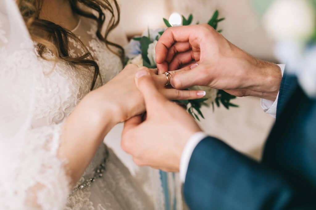 Nahaufnahme eines Bräutigams, der der Braut während der Trauung behutsam einen Ring an den Finger steckt – ein inniger Moment, der oft in einem Eheseminar thematisiert wird. Die Braut trägt ein weißes Spitzenkleid, der Bräutigam einen dunklen Anzug. Im Hintergrund ist Grün zu sehen.
