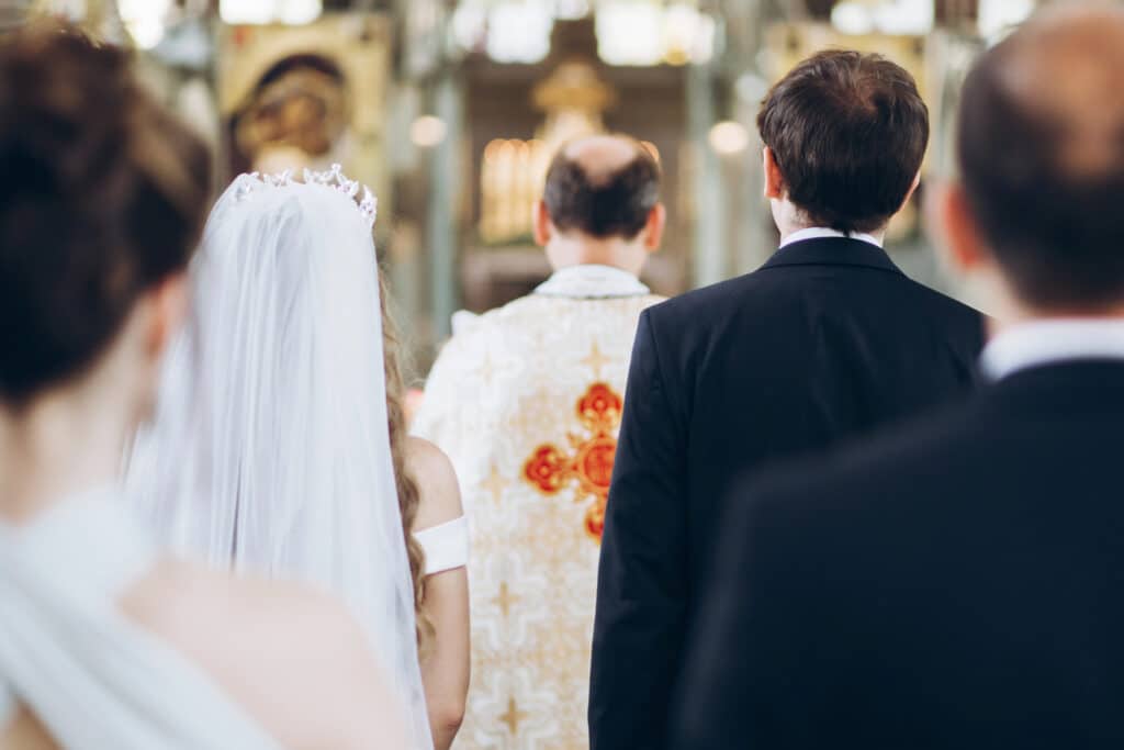 Eine Braut in einem weißen Kleid und mit Schleier steht während einer Trauung neben einem Bräutigam im Anzug, blickt auf einen Priester in der Kirche und ist bereit, nach Abschluss ihres Eheseminars das Eheversprechen zu geben.