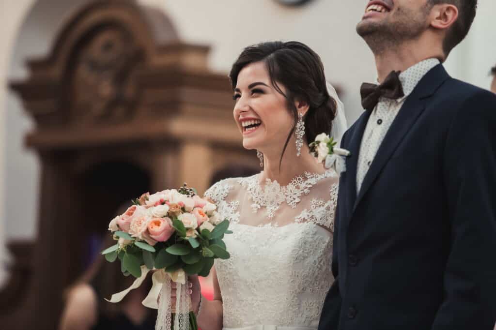 Eine Braut in einem weißen Spitzenkleid, die einen Strauß rosa Rosen in den Händen hält, lächelt fröhlich neben einem Mann in Anzug und Fliege. Beide wirken glücklich, während sie in einem warm beleuchteten Raum stehen und sich vielleicht an schöne Momente aus ihrem Eheseminar erinnern, während sie ihren besonderen Tag feiern.