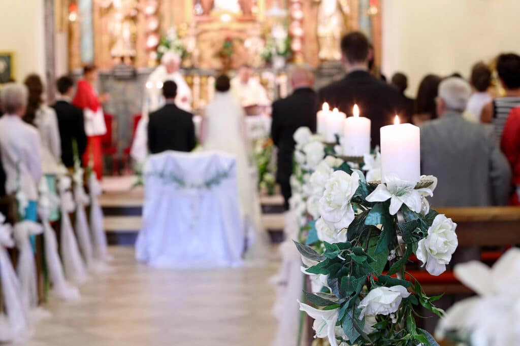 Weiße Kerzen und Blumen schmücken den Kirchengang, auf dem unscharf abgebildete Gäste – vielleicht gerade erst durch ihr Eheseminar inspiriert – sitzen, während im Hintergrund am Altar eine Trauung stattfindet.