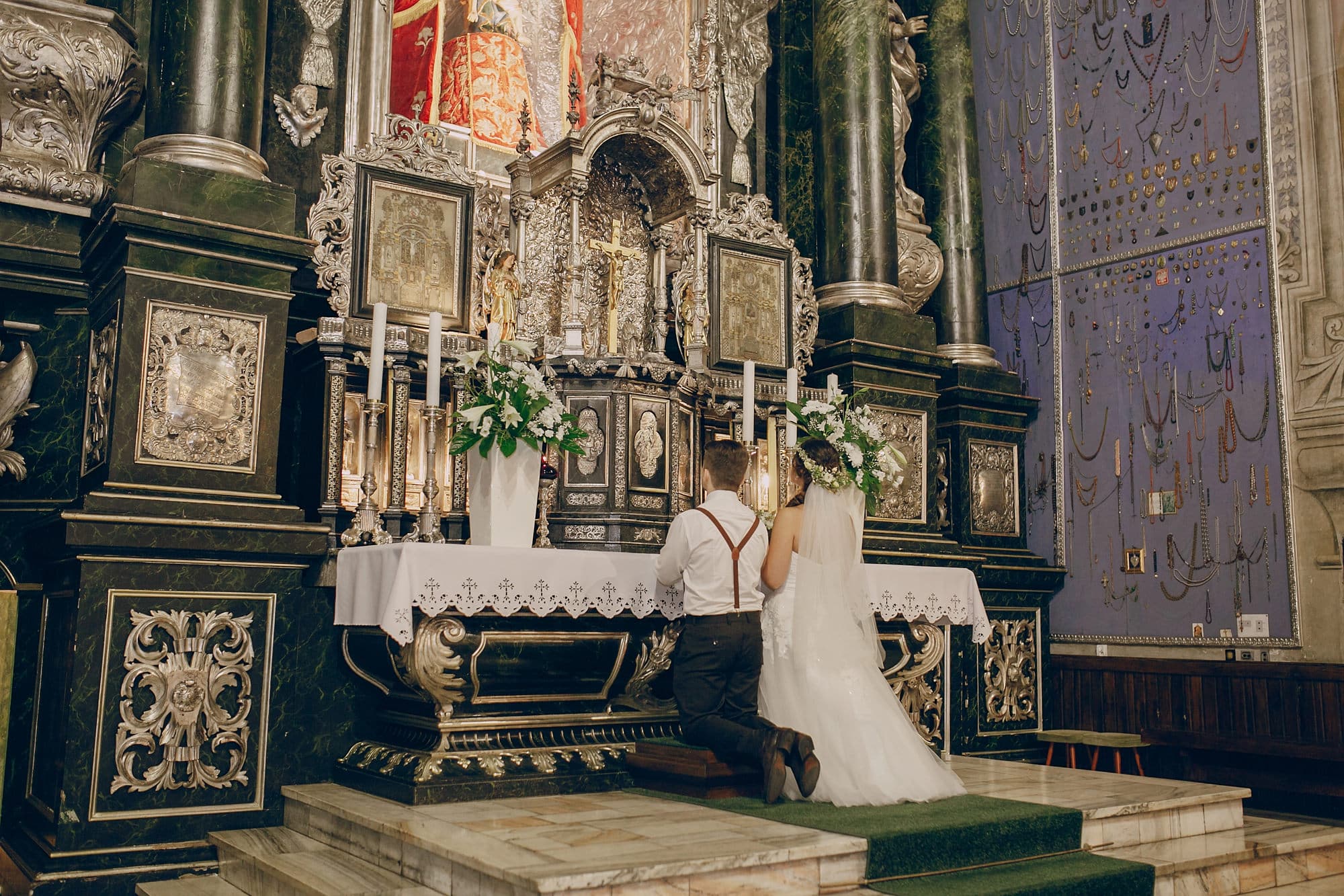 Ein Brautpaar, das gerade sein Eheseminar abgeschlossen hat, kniet gemeinsam vor einem reich verzierten Kirchenaltar, der mit Kerzen und Blumen geschmückt ist, und wendet der Kamera den Rücken zu.