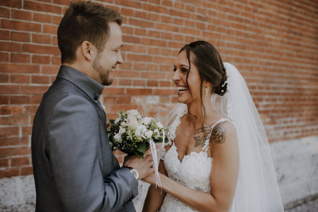 Braut und Bräutigam lächeln sich an ihrem Hochzeitstag an. Die Braut in weißem Kleid und Schleier hält einen Blumenstrauß. Der Bräutigam in einem grauen Anzug steht vor einer Backsteinmauer. Beide wirken fröhlich und unterhalten sich über ihre gemeinsame Hochzeitsreise.