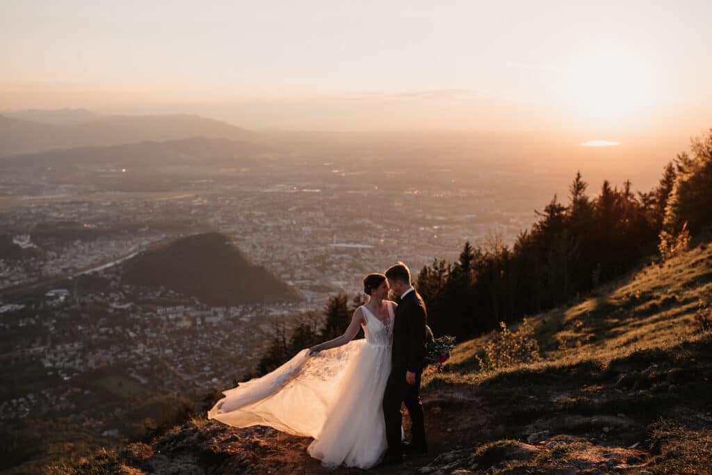 Ein Paar im Hochzeitskleid steht auf einem Berghang, der Schleier der Braut weht im Wind. Umgeben von Bäumen und mit Blick auf eine Stadtlandschaft bei Sonnenuntergang genießen sie die warme, romantische Atmosphäre ihrer Hochzeit.