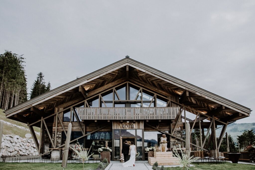 Eine Braut im weißen Brautkleid geht auf ein großes Holzchalet mit dreieckigem Dach und Glasfassade zu, perfekt für jede Hochzeitsplanung. Das Gebäude ist von üppigem Grün und hohen Bäumen unter einem bewölkten Himmel umgeben und schafft eine idyllische Kulisse zum Heiraten.