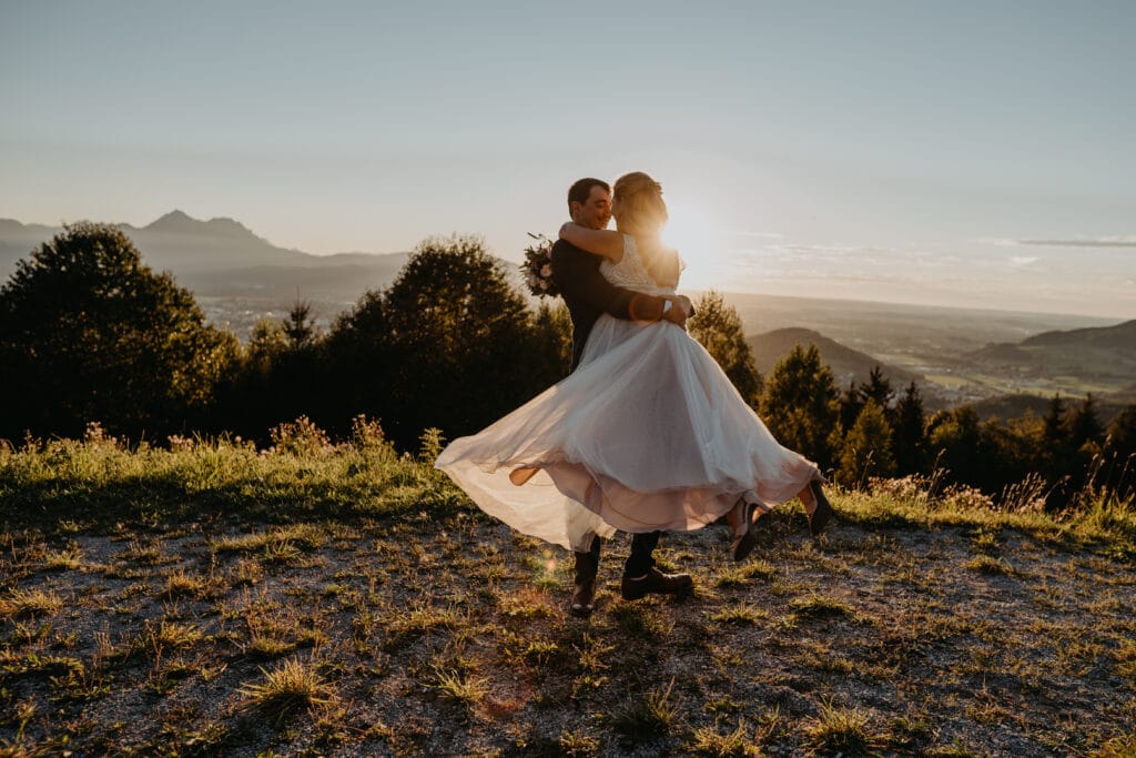 Ein Paar umarmt sich und dreht sich fröhlich auf einem Hügel bei Sonnenuntergang, mit Panoramablick auf Berge und Tal, was eine traumhafte Kulisse für eine romantische Hochzeit schafft. Das Kleid der Frau bauscht sich anmutig, während die Sonne ihren bezaubernden Moment in einen warmen Schein taucht.
