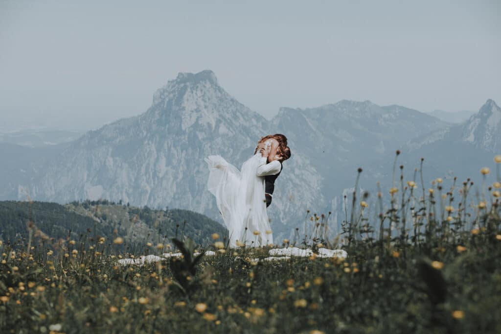 Ein Paar umarmt sich auf einem Berggipfel, umgeben von Wildblumen, während sie von ihrer Hochzeit träumen. Die Kulisse zeigt majestätische Berge unter einem klaren Himmel, die eine heitere und romantische Atmosphäre schaffen, die ihre Heiratspläne inspiriert.