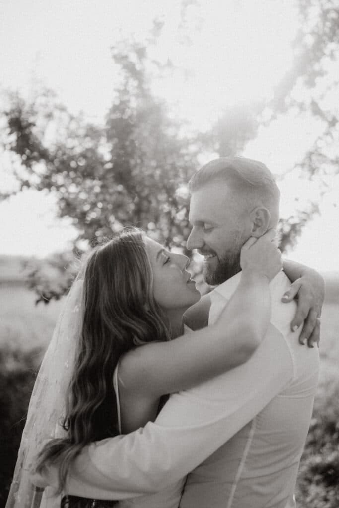 Ein Schwarzweißfoto zeigt ein sich umarmendes Paar im Freien und verkörpert die Essenz von „heiraten“. Die Frau trägt einen Schleier und lange Haare, der Mann einen kurzen Bart. Sie blicken einander in die Augen, eingerahmt von Bäumen und Sonnenlicht – ein zeitloser Moment der Liebe inmitten der Schönheit der Natur.