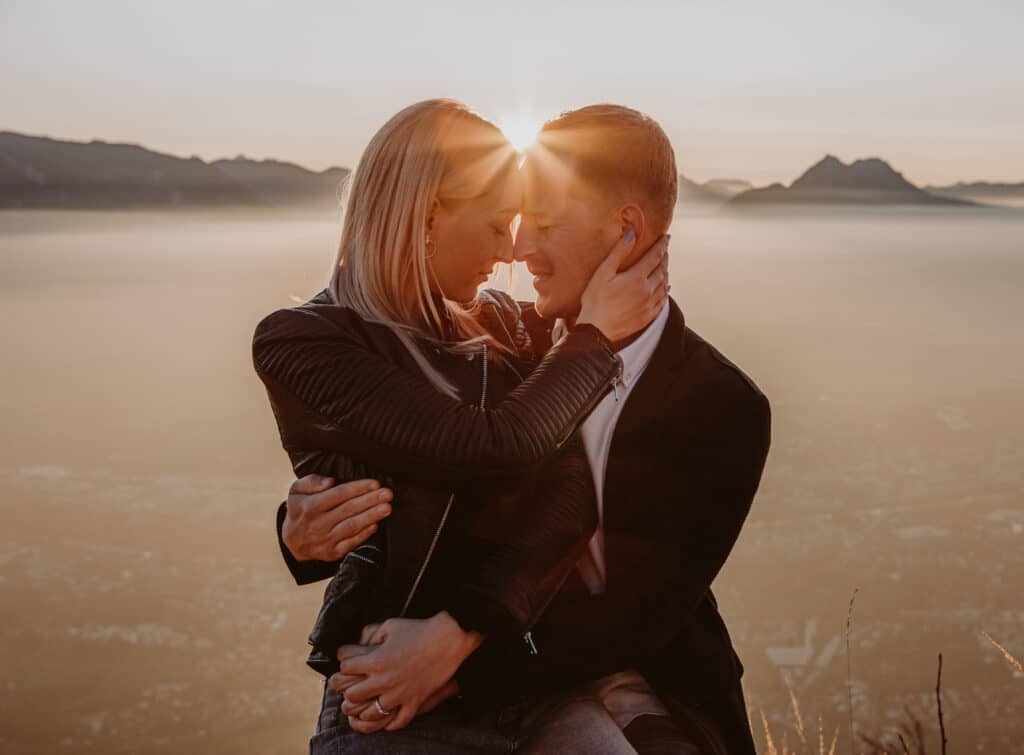 Ein Paar umarmt sich bei Sonnenaufgang auf einem Berggipfel, ihre Stirnen berühren sich, getaucht in warmes Sonnenlicht. Inmitten einer dunstigen Skyline und Berggipfeln erleben sie einen romantischen und ruhigen Moment, der ihre Hochzeitspläne andeutet.